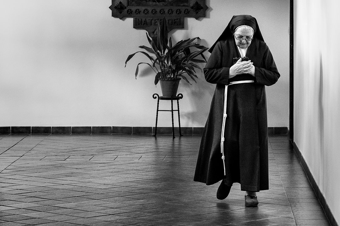 A nun with her hands clasped beside a crucifix on the wall.